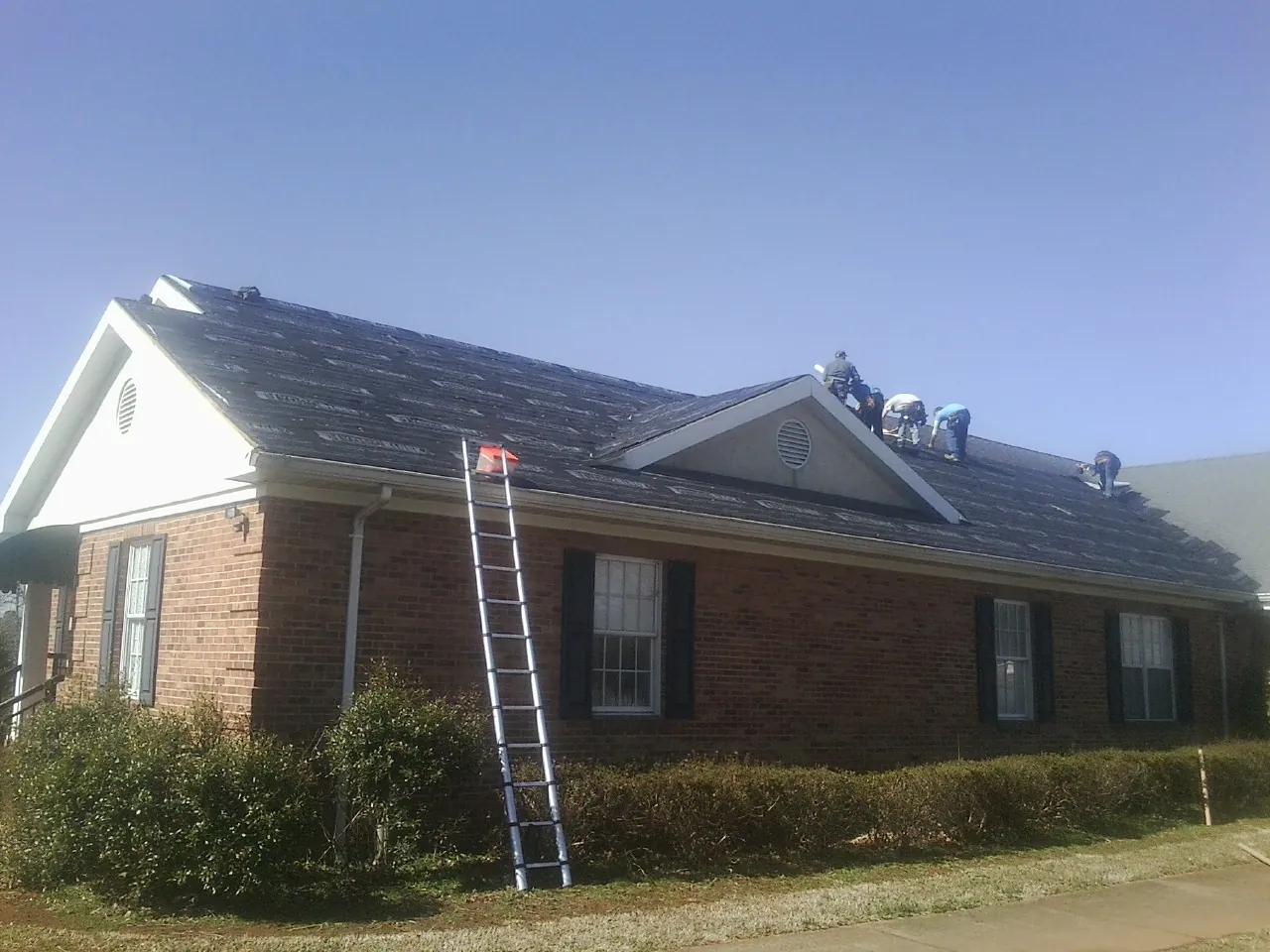Crew tearing shingles off a brick ranch roof with a ladder set — Metal Roof Repair work by our crew in Robins