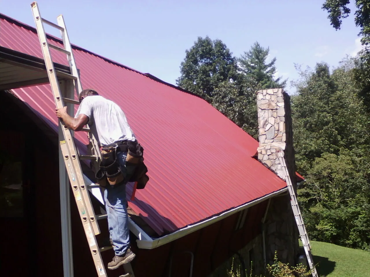 Roofer on a ladder beside a red metal roof and stone chimney — Soffit & Fascia Repair by Roofing Brothers in Robins